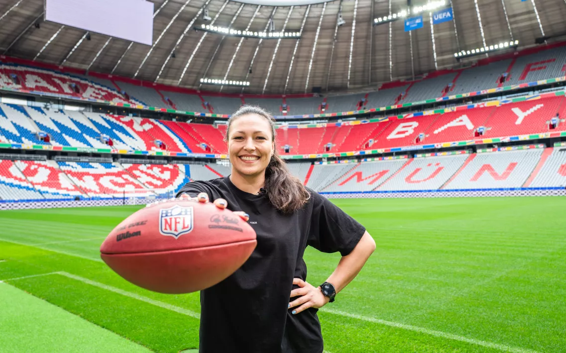 Nadine Nurasyid steht neben dem Rasen in der Allianz Arena und hält einen Football in der rechten Hand