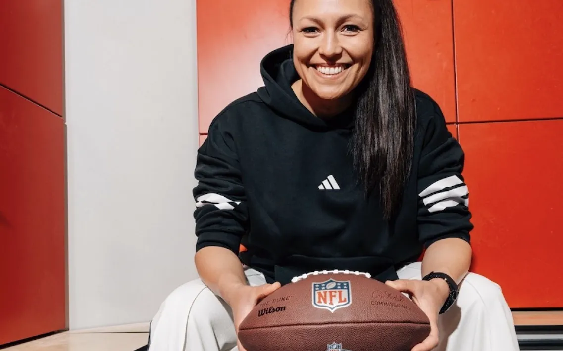Nadine Nurasyid sitzt mit einem Football in der Hand auf einer Bank in einer Umkleidekabine